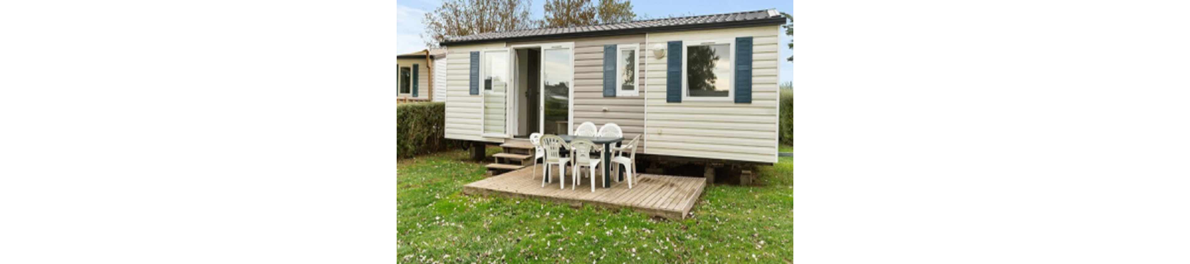 A modern mobile home is pictured with a small wooden patio at the entrance. The patio features a round table and several white plastic chairs, set on a grassy lawn with scattered leaves. The home has blue shutters, a large window, and a glass door, with trees and hedges in the background. The scene suggests a relaxed, outdoor setting suitable for gatherings. No visible text or numbers are present.