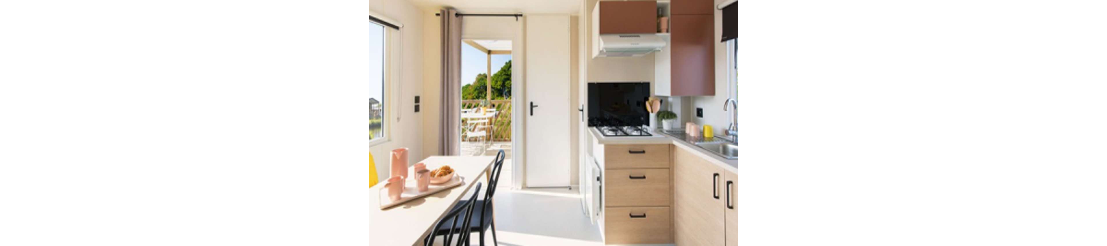 A bright, modern kitchen and dining space features light wood cabinetry, a gas stove, and a small dining table set for breakfast. Sunlight streams in through large windows and a glass door, revealing an outdoor patio with greenery. The interior is decorated with pastel accents, including pink and yellow tableware, creating a fresh and inviting atmosphere.