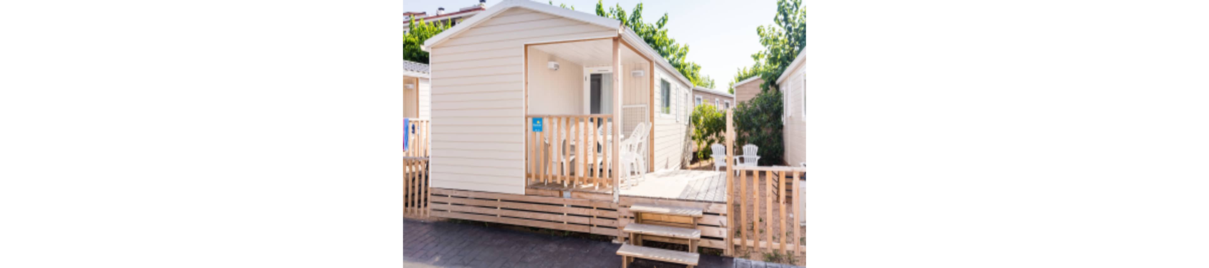 A light-colored mobile home features a small wooden porch with white chairs and a table. The setting is outdoors, surrounded by greenery and neighboring similar units. Two white Adirondack chairs are visible on the sandy area next to the porch, creating a relaxed and inviting atmosphere.