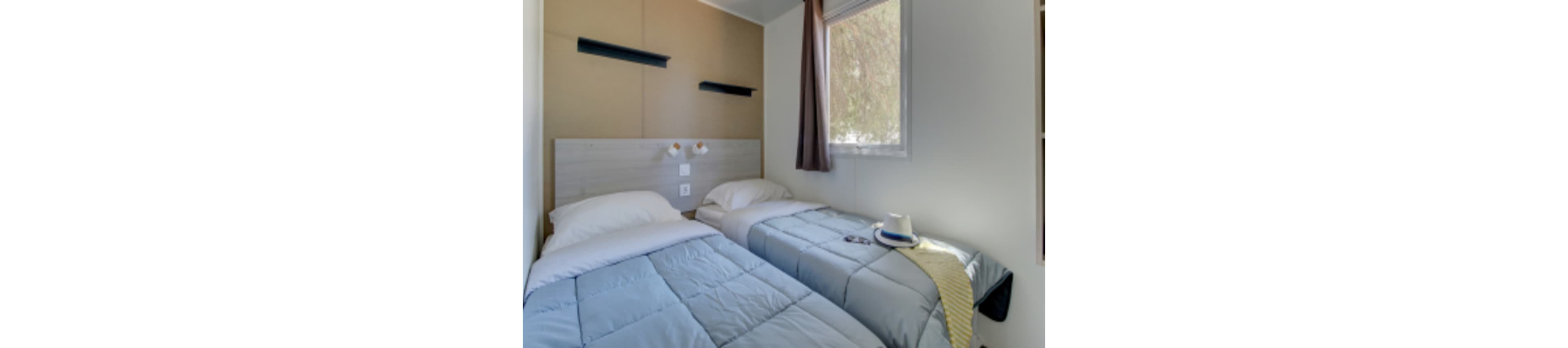 A small bedroom features two neatly made twin beds with light blue comforters and white pillows. A wooden headboard with two wall-mounted reading lights and shelves adds simple, modern style. Daylight filters through a window with a brown curtain, creating a calm atmosphere. A hat, belt, and phone on the bed suggest a relaxed, vacation-like setting.