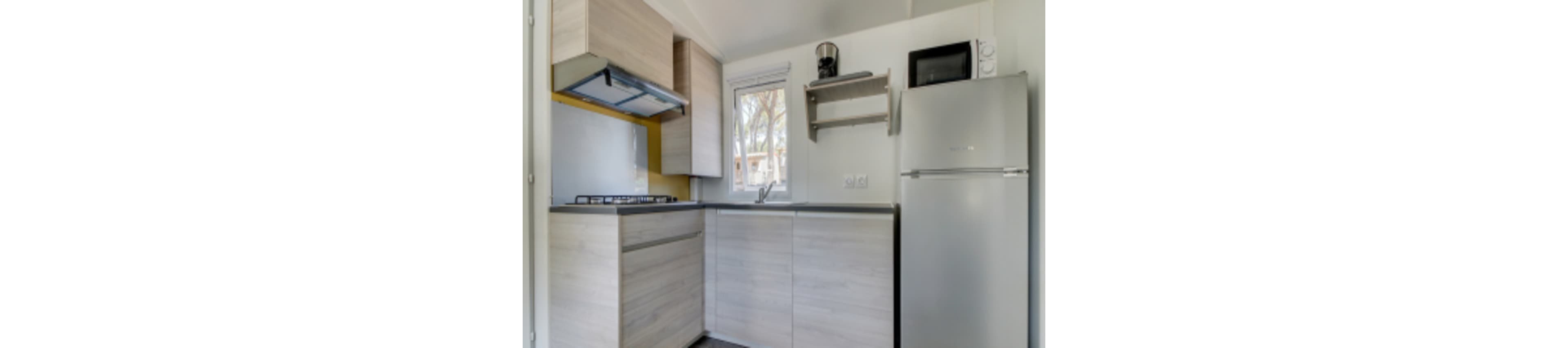 A small, modern kitchen features light wood cabinetry and a sleek countertop. The space includes a refrigerator, microwave, and a gas stove with a range hood. Natural light enters through a window above the sink, and open shelving holds a coffee maker. The overall palette is neutral with clean lines and minimal decor.