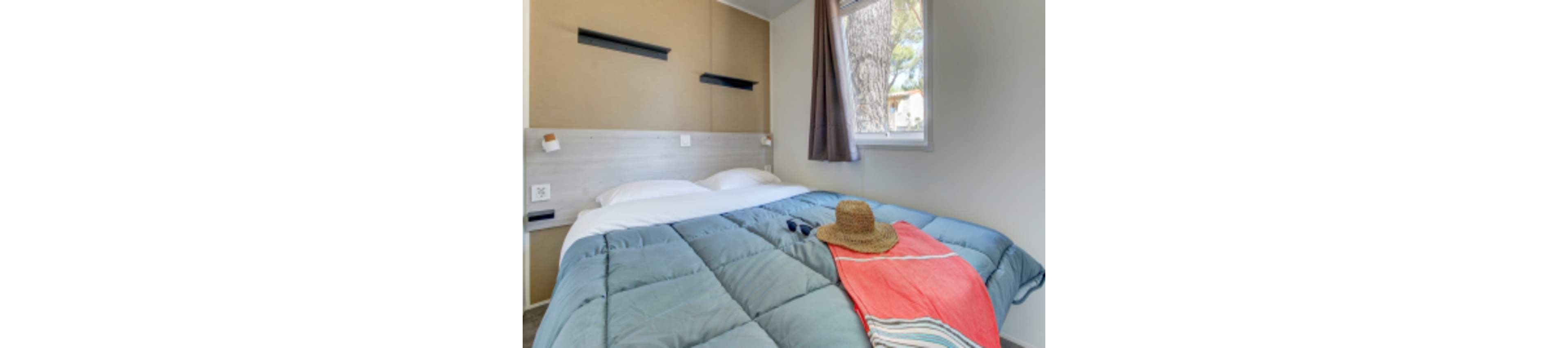 A neatly made bed with a blue comforter is the main focus in this modern, compact bedroom. A straw hat, sunglasses, and a coral towel are placed on the bed, suggesting a relaxed, vacation atmosphere. The room features neutral walls, a window with a view of trees, and minimalistic decor, creating a calm and inviting mood.