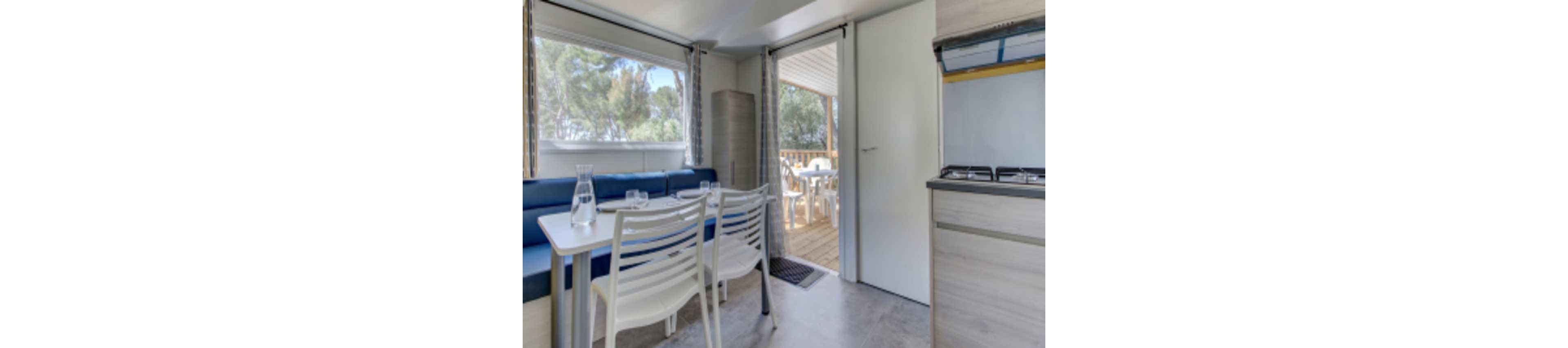A compact kitchen and dining space is shown inside a modern mobile home. The area features a small table with white chairs, a blue cushioned bench, and a kitchenette with a stove. Large windows and an open door reveal a wooden deck with outdoor seating and trees in the background. The setting is bright and airy, with a relaxed vacation atmosphere.