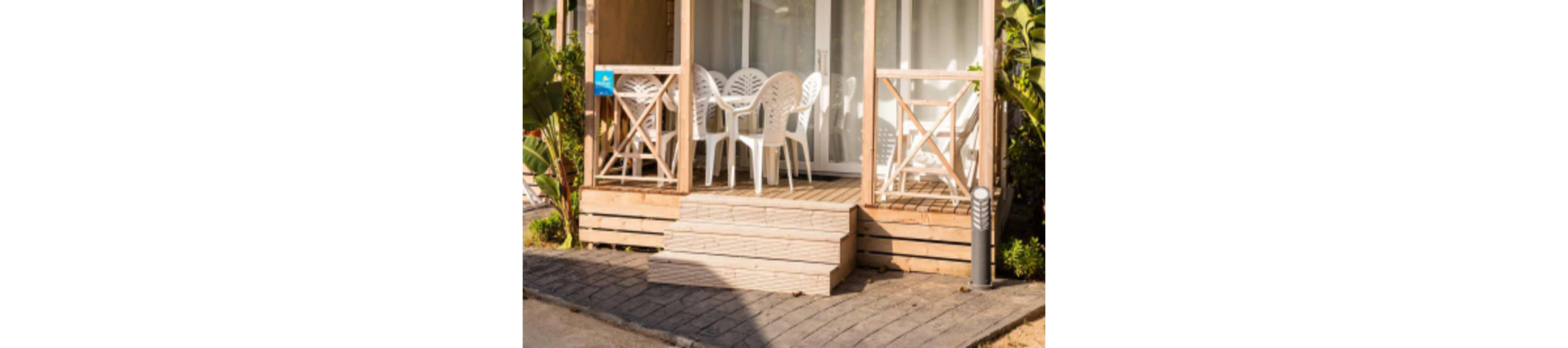 A small wooden porch features several white plastic chairs and a matching table, set in front of a glass door. The area is bathed in sunlight, creating a warm and inviting atmosphere. Lush green plants frame the porch, and a paved walkway leads up to the steps. The overall mood is relaxed and summery, ideal for outdoor leisure.