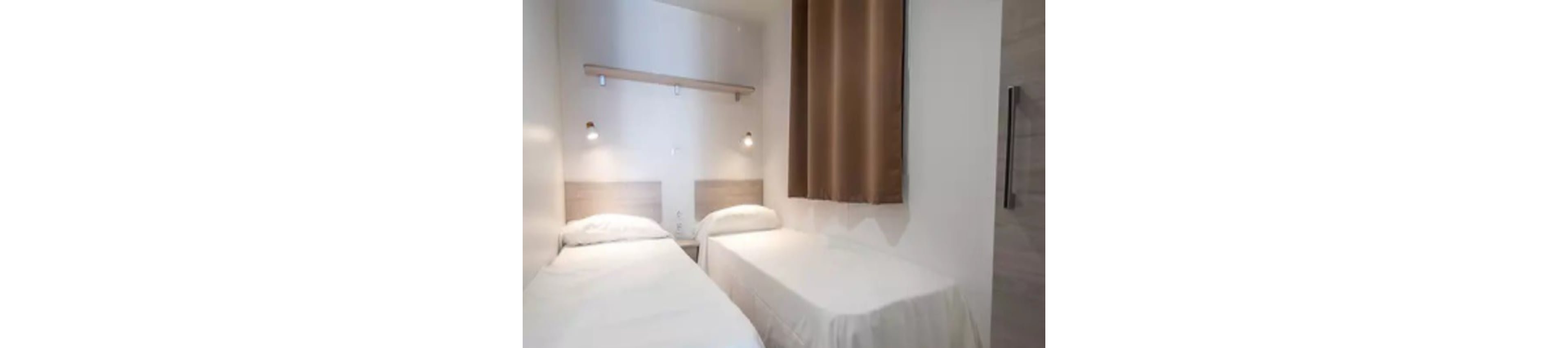 This image shows a compact hotel room featuring two single beds with white linens and light wood headboards. The setting is simple and modern, with wall-mounted reading lights above each bed and a brown curtain covering the window. The overall palette is neutral, dominated by whites and light browns, creating a clean and calm atmosphere. No visible text or numbers are present in the image.