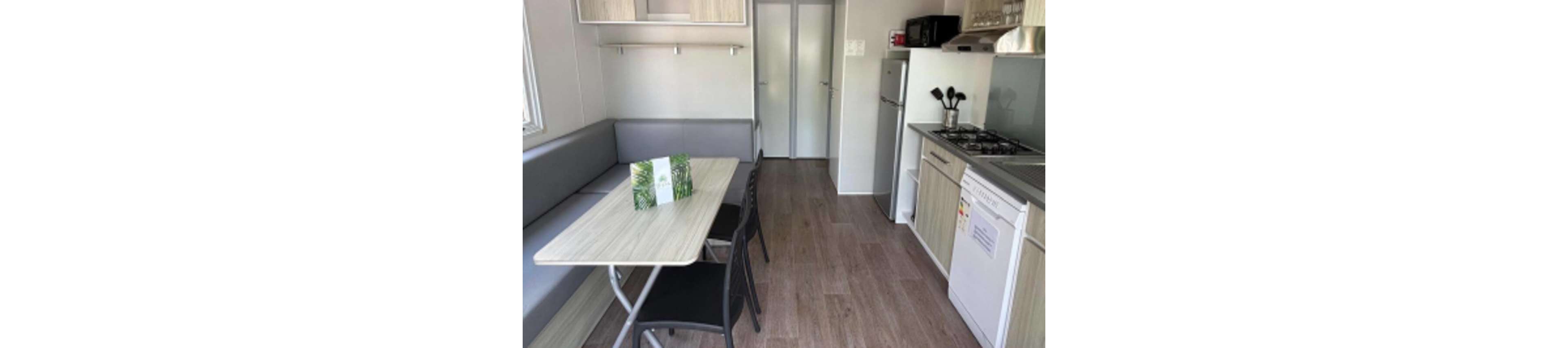 A small, modern kitchen and dining space features a built-in bench, table, and two chairs. The kitchen includes a refrigerator, microwave, stove, and utensils, with light wood cabinetry and neutral tones. A folded brochure or menu sits on the table, and the room is well-lit with natural light from a window. The overall style is minimalist and functional, suitable for a compact living environment.