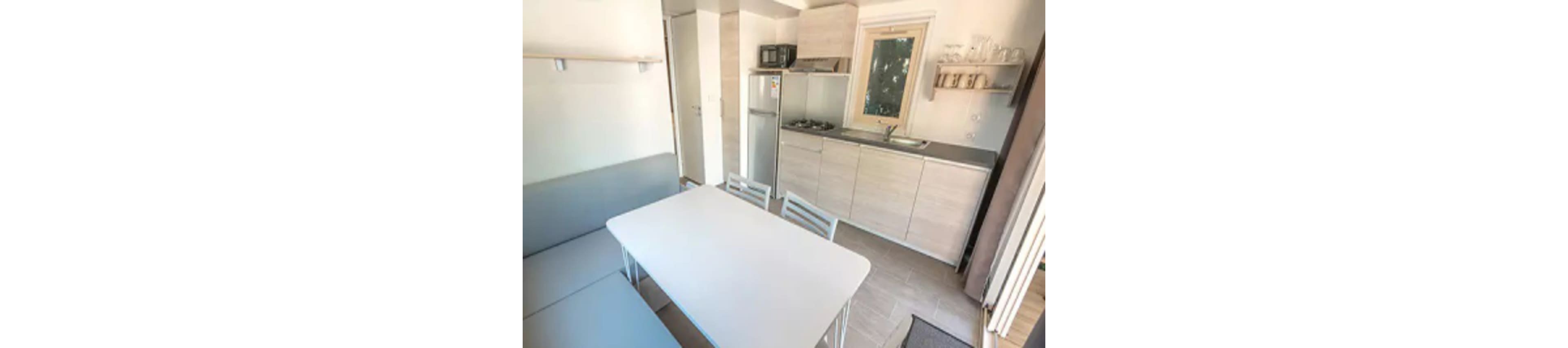A small, modern kitchen is shown with light wood cabinetry, a compact refrigerator, and a microwave. The dining area features a white table with metal chairs and a built-in bench. The space is bright with neutral tones, minimal decor, and open shelving displaying glassware. Natural light enters through a window, creating a clean and inviting atmosphere.