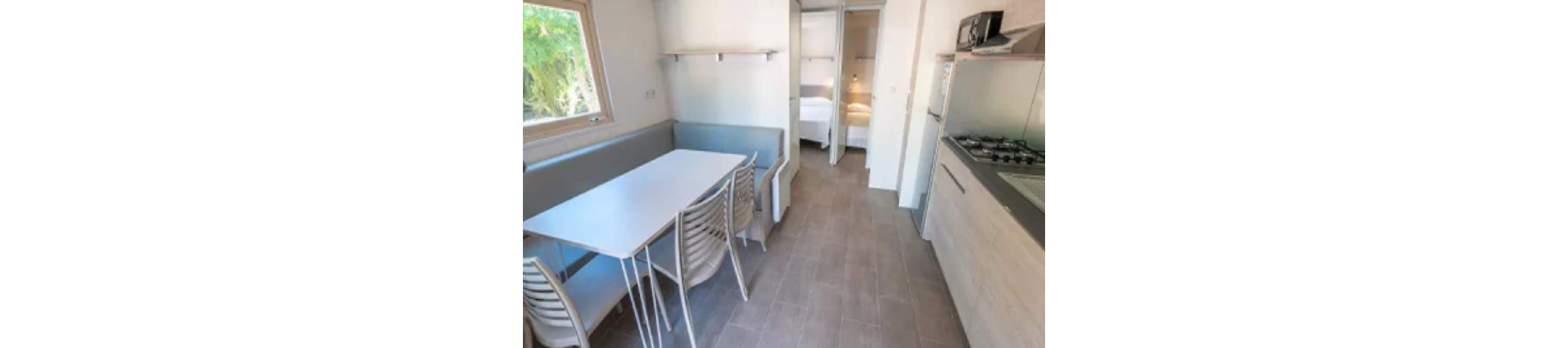 A bright, modern kitchen and dining space features a sleek table with four chairs and a built-in bench. The room is accented by neutral tones and natural light from a window, with a view into two adjoining bedrooms. The kitchen includes a gas stove, microwave, and minimalist cabinetry, creating a clean and functional atmosphere.