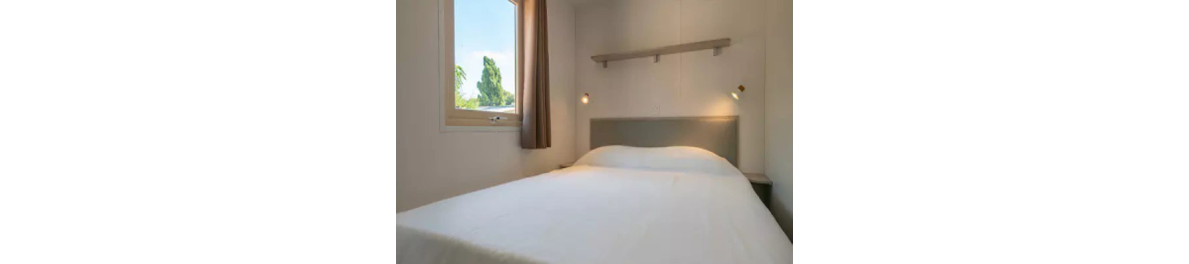 A simple, modern bedroom features a neatly made bed with a white comforter and a gray headboard. The room includes a window with brown curtains, allowing natural light to brighten the space. A floating wall shelf and two wall-mounted reading lights add functional decor. The overall palette is neutral, creating a calm and inviting atmosphere.
