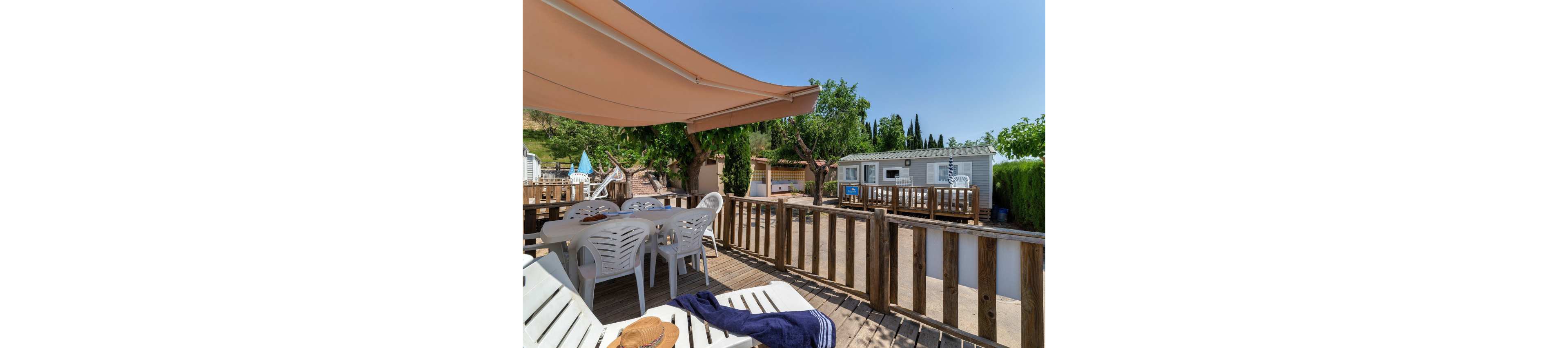 A wooden terrace with white plastic chairs and loungers sits in front of a mobile home under a large sunshade. The scene is set on a bright sunny day with clear blue sky and surrounding greenery. A straw hat and dark blue towel rest on a lounger, suggesting a relaxed vacation atmosphere. Other mobile homes and trees are visible in the background, creating a cozy holiday park setting.
