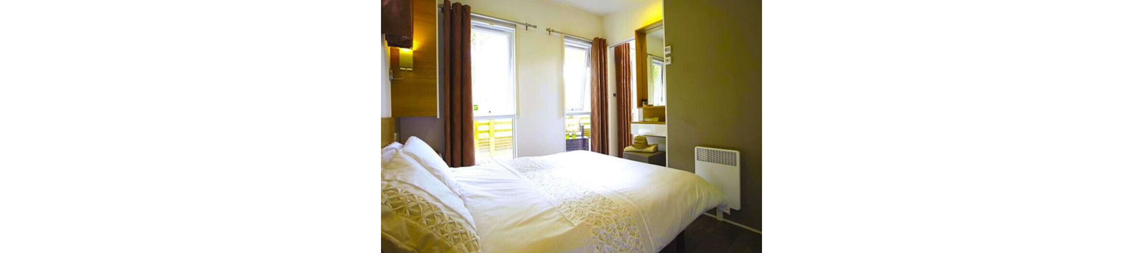 A neatly made bed with white linens is the focal point of this modern bedroom. The room features two large windows with brown curtains, allowing natural light to fill the space. A wall-mounted mirror and a small vanity area are visible, along with a compact white heater. The overall palette is neutral with warm accents, creating a cozy and inviting atmosphere.