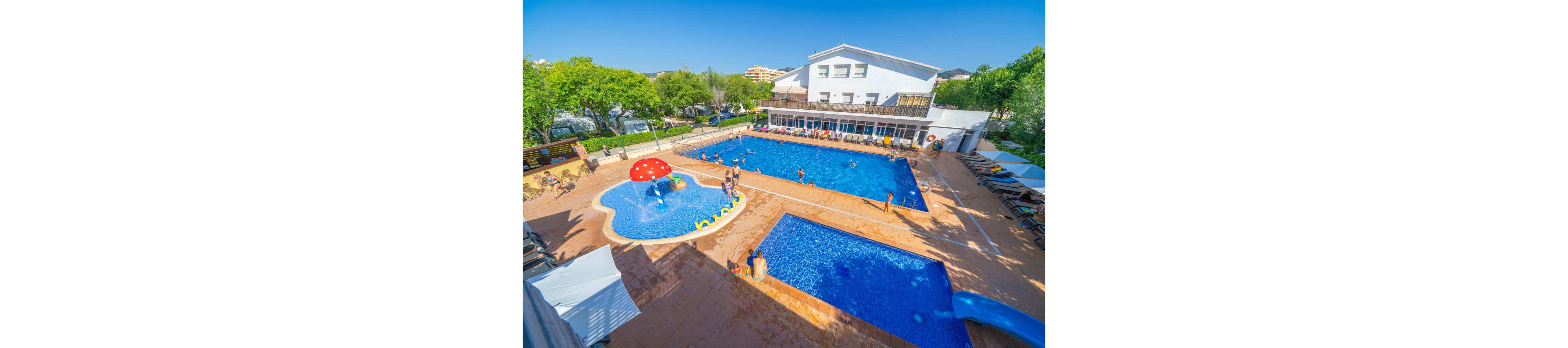 A bright family resort features multiple blue outdoor swimming pools beside a white hotel building. Children and adults enjoy the water, with kids playing in a shallow pool that has a red mushroom fountain and small slides. Sun loungers line the pool deck under clear blue skies, surrounded by green trees. The overall scene suggests a warm, relaxing vacation atmosphere at a family-friendly hotel.