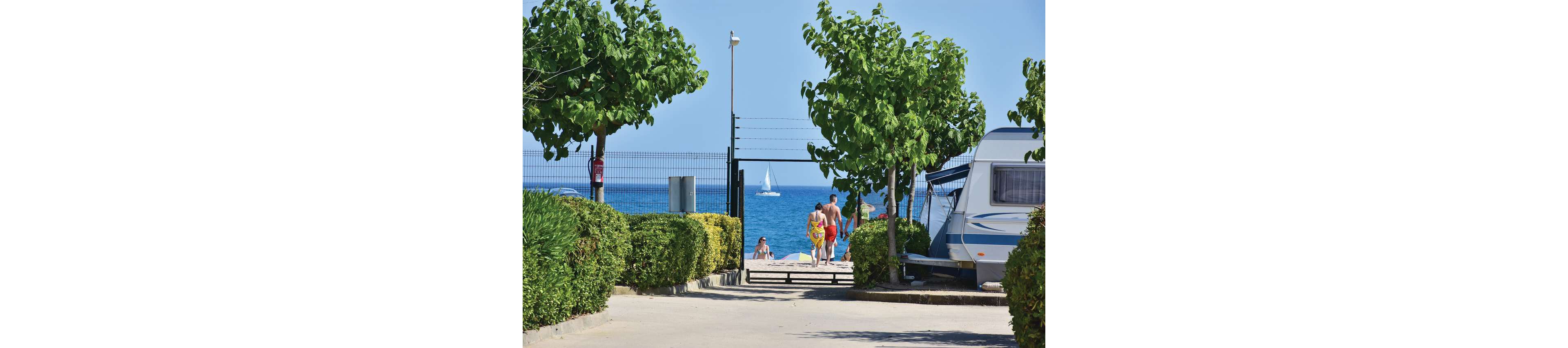 A family in swimwear walks from a campsite pathway toward a bright sandy beach. A caravan is parked on the right, framed by neatly trimmed hedges and small trees. Beyond the fence, people relax near the shoreline while a white sailboat glides across the blue sea. The scene suggests a relaxed summer holiday by the coast.