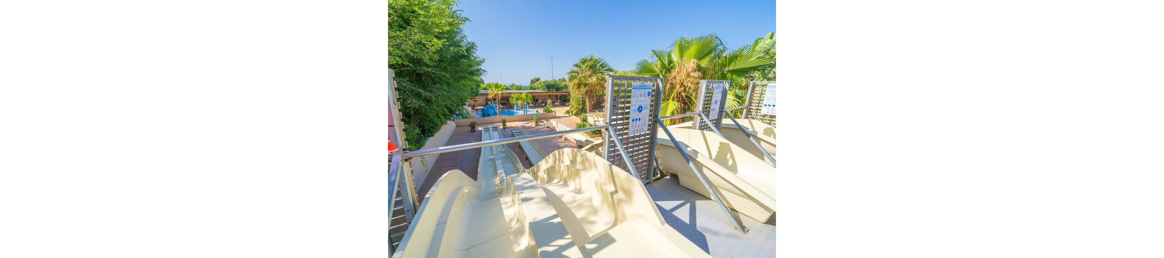 Image of several light-colored water slides leading down toward an outdoor pool area. The setting appears to be a sunny water park surrounded by palm trees and lush greenery. Metal railings and safety instruction boards are visible near the slide entrances. In the background, people relax near the pool under clear blue skies, creating a bright and inviting vacation atmosphere.
