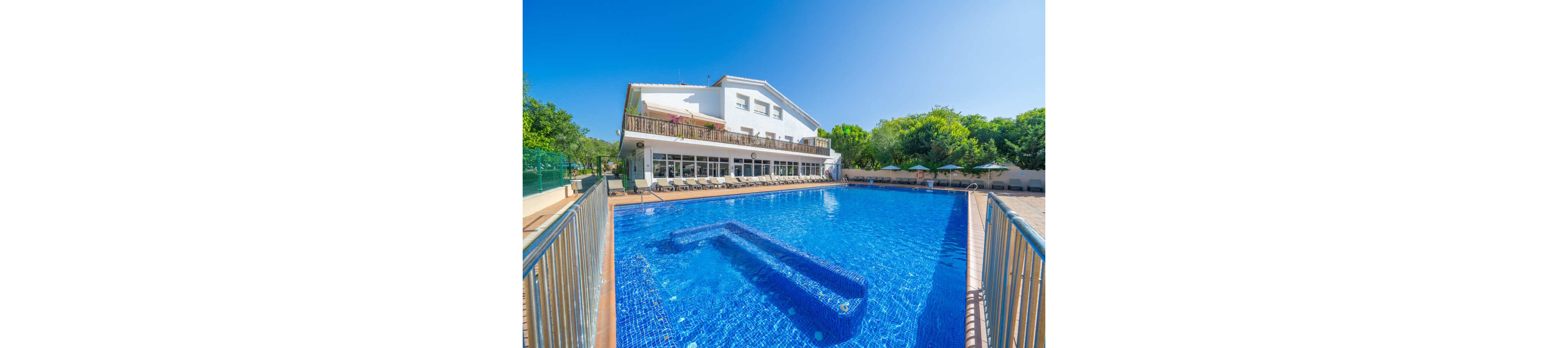 A large outdoor swimming pool dominates the foreground, its clear blue water reflecting the bright sky. Behind the pool stands a modern white hotel building with balconies and large windows. Sun loungers and umbrellas line the poolside terrace, suggesting a relaxing vacation setting. Lush green trees surround the area, adding a natural backdrop to the sunny, tranquil scene.