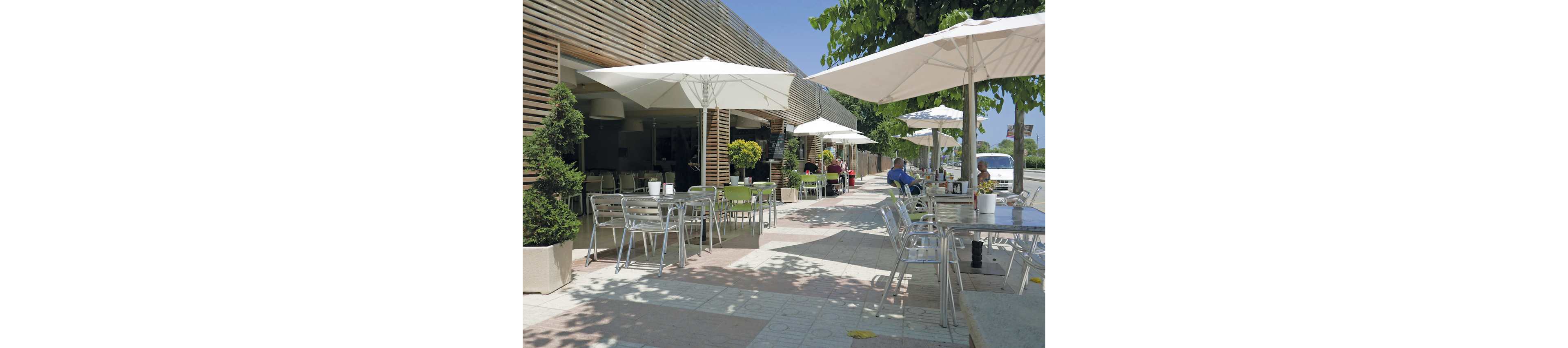 An outdoor café terrace features rows of metal tables and chairs arranged along a wide sidewalk. Large white umbrellas provide shade for diners seated near the building and under nearby trees. The modern façade with wooden slats contrasts with the greenery of potted plants and street trees. A few people sit and relax at the tables, creating a casual, sunny atmosphere in an urban setting.