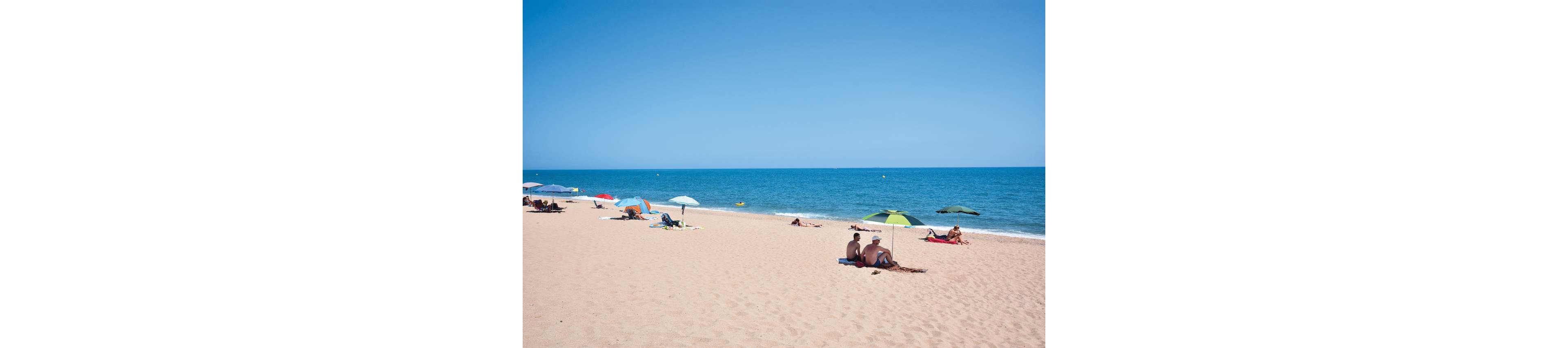A wide sandy beach stretches along a calm blue sea under a clear sky. Small groups of people relax on towels and loungers, some sitting under colorful umbrellas for shade. The shoreline is gently lapped by waves, and the open water extends to the horizon. The overall scene feels peaceful, spacious, and summery.