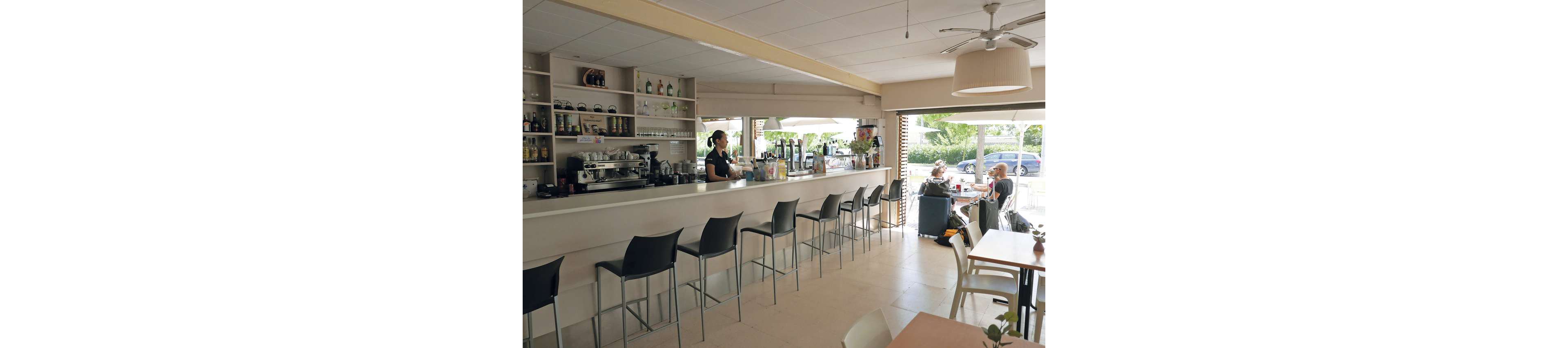 A modern cafe interior features a long white counter with a barista preparing drinks behind it. Shelves stocked with bottles, cups, and a large espresso machine line the back wall. Several empty bar stools face the counter, while customers sit at low tables near large open windows. Natural light fills the space, highlighting the clean, minimalist decor and relaxed atmosphere.