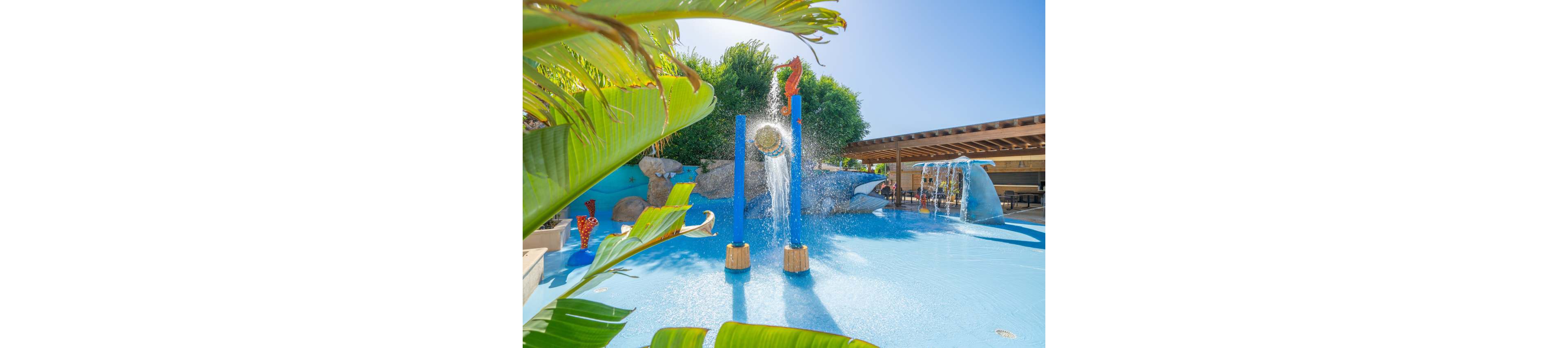An outdoor children’s splash park features playful blue pillars spraying water into a shallow pool. A seahorse figure and other colorful aquatic decorations create a fun, themed environment. The setting is bright and sunny with clear blue skies and lush green plants framing the scene. A shaded structure and additional water features are visible in the background, suggesting a family-friendly resort or water park area.