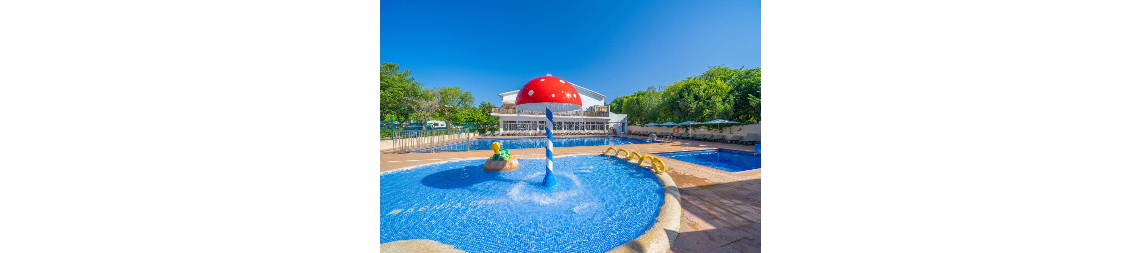 A vibrant children’s splash pad features a large red mushroom-shaped water fountain in the foreground. The shallow circular pool includes playful animal water features and curved yellow structures for kids to climb and splash around. Behind the splash area, a larger swimming pool and a modern resort building are visible. Lush green trees and a clear blue sky create a bright, sunny holiday atmosphere.