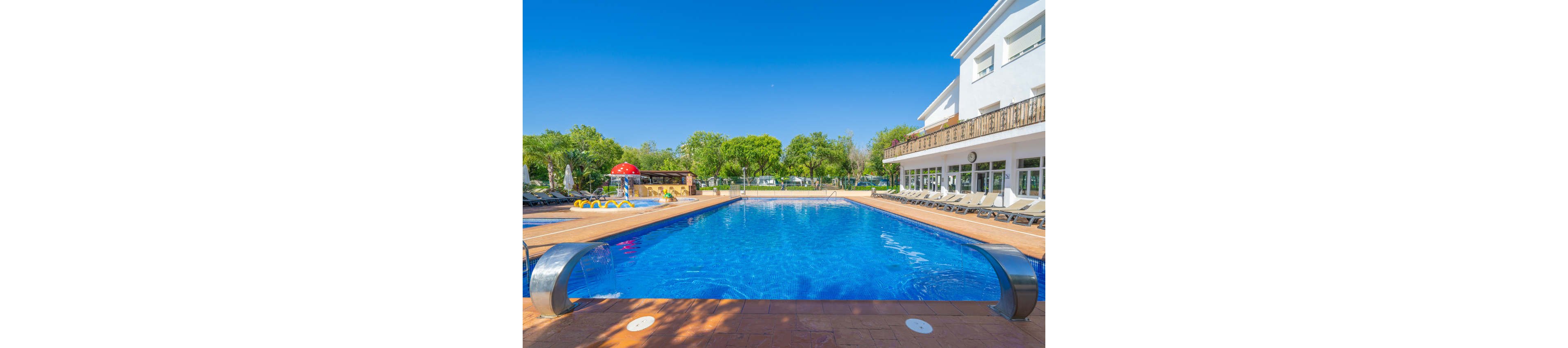 A large outdoor swimming pool glistens under a clear blue sky at a resort or hotel. Sun loungers line the poolside terrace next to a white building with large windows and balconies. In the background, a small children's water play area with slides and a mushroom fountain adds a playful touch. Lush green trees surround the scene, creating a relaxing vacation atmosphere.