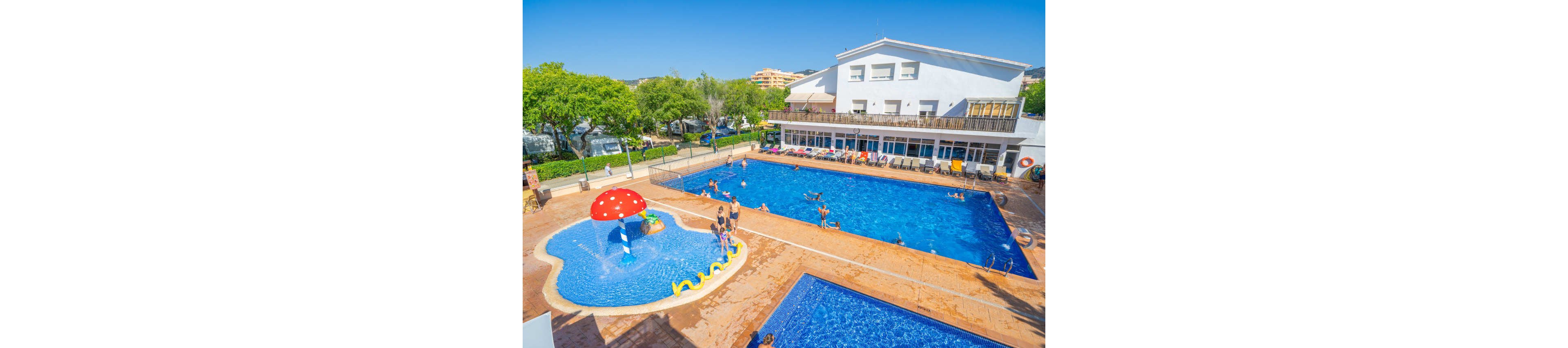 A bright family resort features multiple outdoor swimming pools beside a white two-story building. Children and adults enjoy swimming and playing in the water under clear blue skies. A shallow kids’ pool includes a red mushroom-shaped fountain and a yellow snake water feature. Green trees and distant buildings frame the leisure area, creating a relaxed holiday atmosphere.