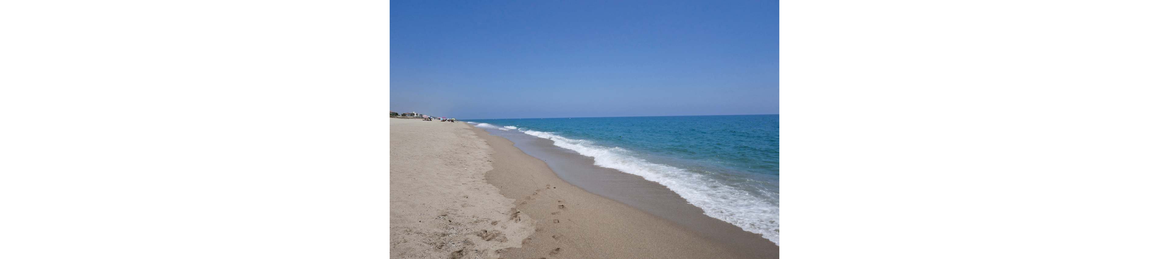 A quiet sandy beach stretches into the distance beside a calm blue sea. Gentle waves roll onto the shore under a clear, cloudless sky. A line of footprints leads along the wet sand near the water’s edge. The scene feels peaceful and remote, with only a few distant figures barely visible.