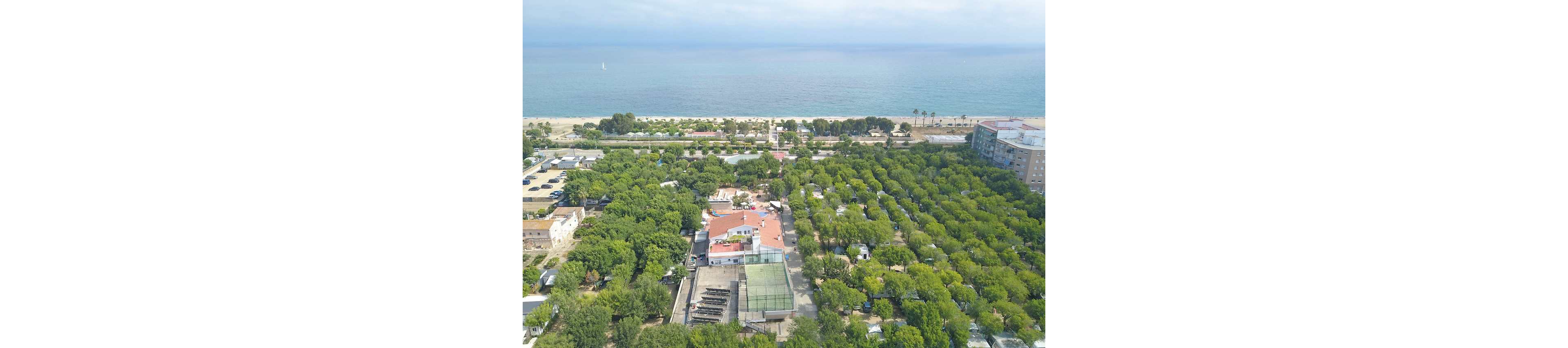 Aerial view of a large campground surrounded by dense green trees near the coastline. Rows of camping plots and small structures are visible among the foliage, with a central building complex featuring red roofs. A tennis or sports court appears in the middle of the site, and nearby urban buildings border one side. In the background, a sandy beach and calm sea stretch to the horizon under a lightly clouded sky.