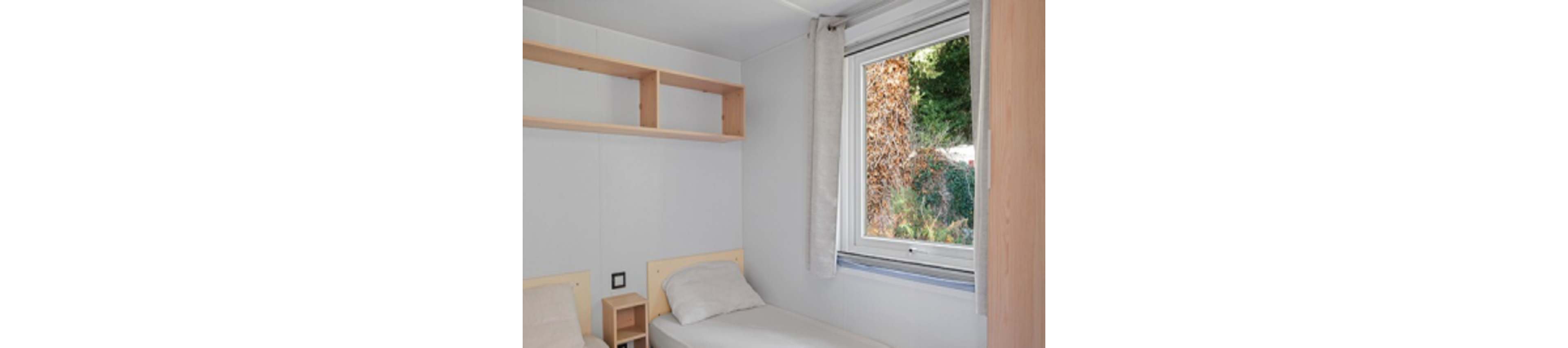 A small, minimalist bedroom features two single beds with light-colored headboards and plain bedding. A simple wooden shelf and compact bedside unit add subtle storage against the white walls. A large window with a neutral curtain lets in natural light and offers a view of greenery and foliage outside. The overall scene feels clean, bright, and functional, suitable for a guest room or vacation rental.