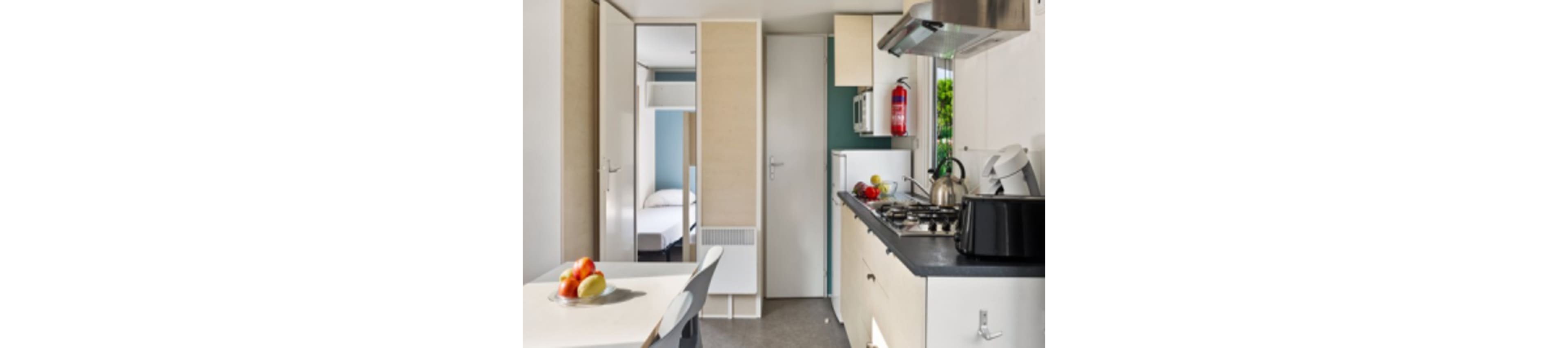 A compact modern kitchenette features a gas stove, sink, and countertop appliances in a small apartment or studio. Light wood cabinets and white walls create a bright, minimalist look. A small dining table with fruit sits in the foreground, while a bedroom is visible through an open doorway. A fire extinguisher and window add functional and natural light elements to the space.