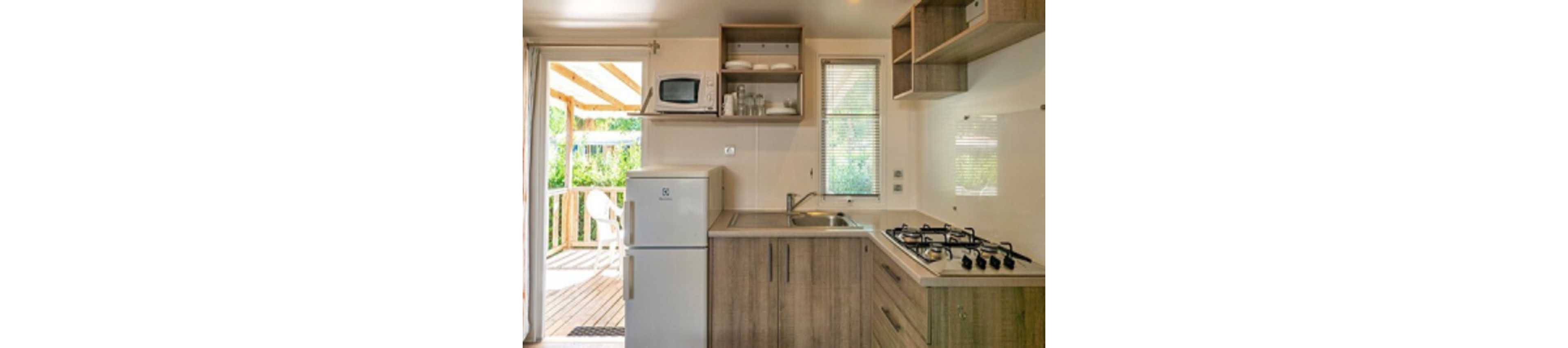 A small modern kitchen features light wood cabinets, a white refrigerator, and a built-in gas cooktop. Open shelving holds dishes and glasses, with a microwave placed above the fridge. A single-basin sink sits beneath a narrow window that lets in natural light. A glass door opens to a sunny wooden deck with outdoor seating, connecting the indoor kitchen to the greenery outside.