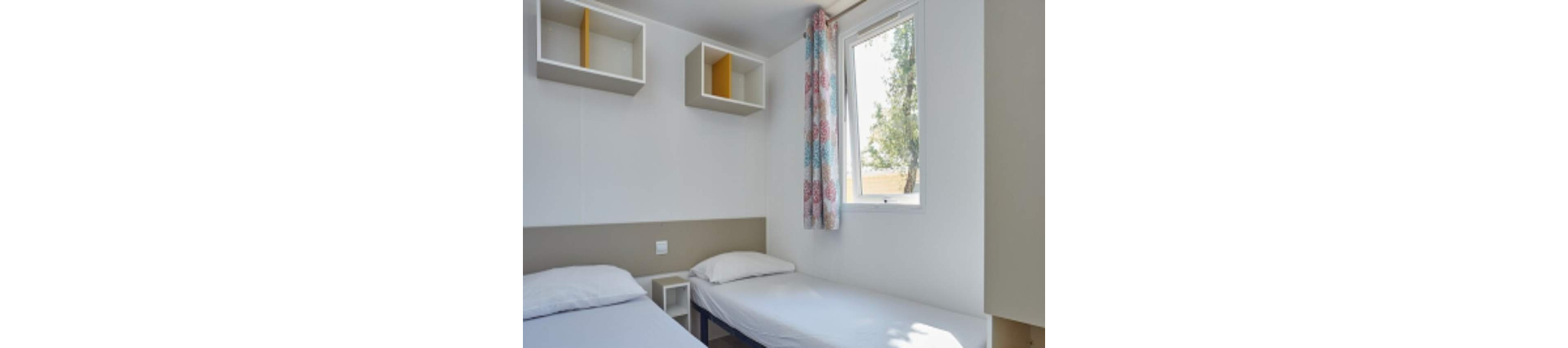 A compact bedroom features two simple twin beds with white bedding against light-colored walls. Above one bed, open cube shelves provide minimal storage and a touch of modern design. A small bedside unit sits between the beds, emphasizing the room’s functional layout. Natural light enters through a window with patterned curtains, creating a bright and airy atmosphere.