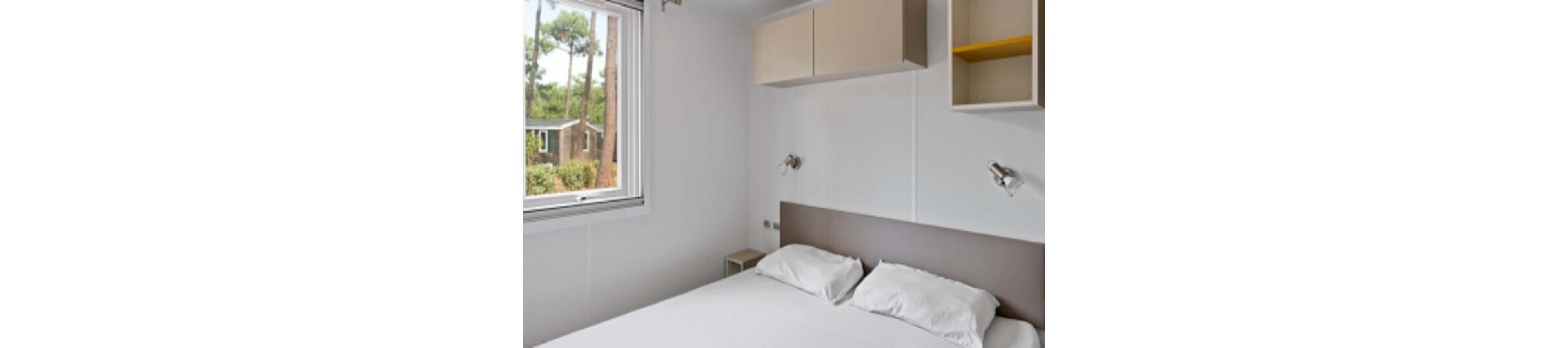 A small minimalist bedroom features a neatly made double bed with white linens. Built-in wall cabinets and open shelving provide storage above the headboard. Two modern reading lights are mounted on the wall on either side of the bed. A window lets in natural light and offers a view of trees and a neighboring building outside.