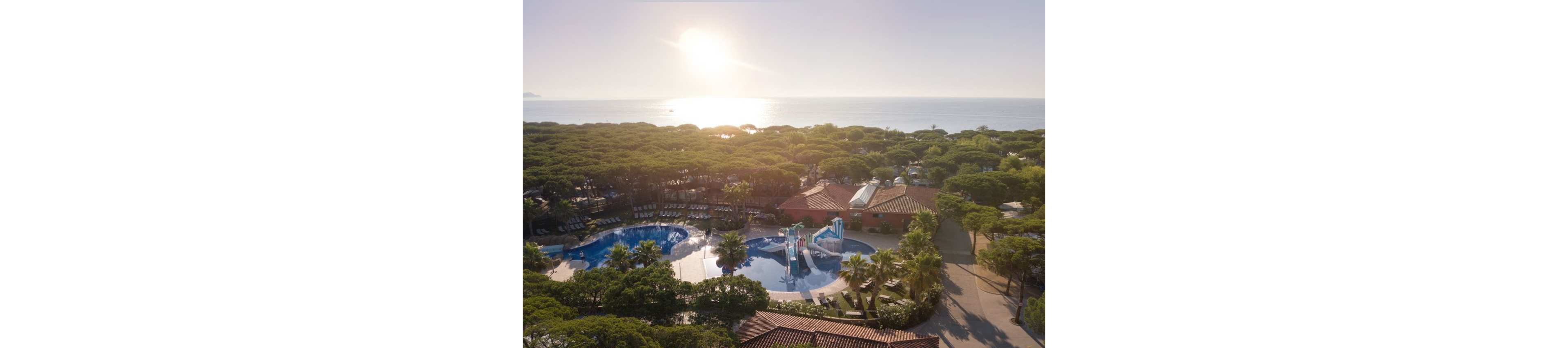 An aerial view shows a coastal resort surrounded by dense green trees near the sea. In the foreground, a large outdoor swimming pool with water slides and play structures is visible. Sun loungers are arranged around the pool area, and low-rise buildings with red-tiled roofs sit among the trees. The sun is low over the horizon, casting a warm glow across the water and landscape.