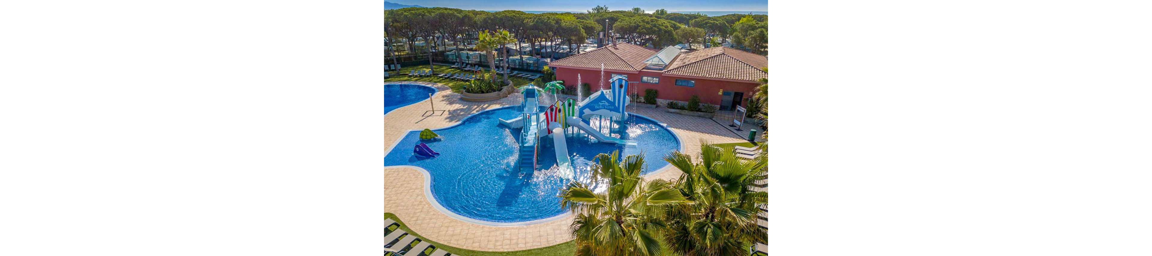 An outdoor resort pool features a colorful water play structure with multiple slides and fountains. The pool is surrounded by tiled walkways, sun loungers, and lush palm trees. In the background, red-roofed buildings and tall pine trees frame the leisure area under a clear blue sky. The scene suggests a family-friendly vacation destination with warm, sunny weather.