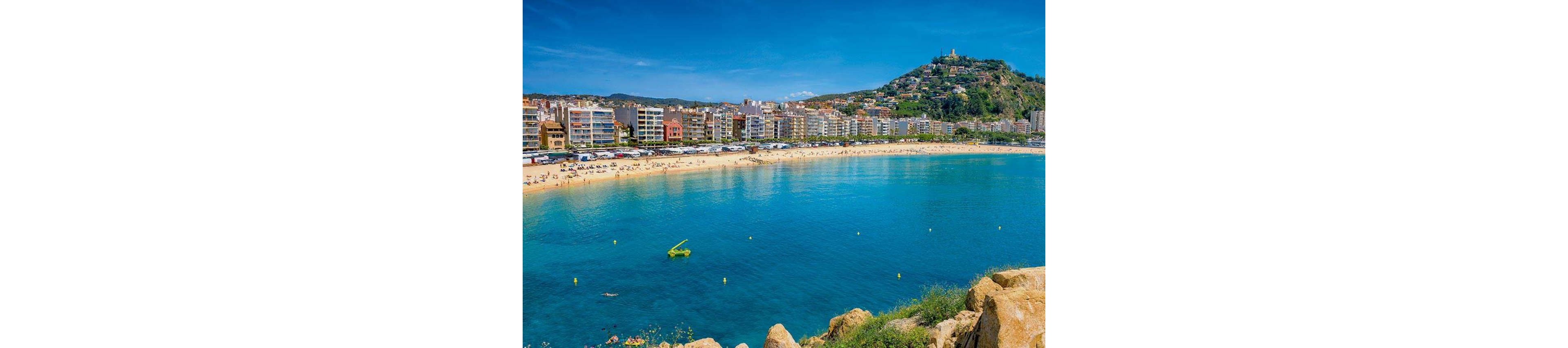 A wide sandy beach curves along a bright turquoise bay in front of a dense line of seaside apartment buildings. Sunbathers and swimmers enjoy the clear water while small yellow buoys float offshore. In the background, a green hill rises above the town with scattered houses and a structure at the summit. The scene is captured under a vivid blue sky, evoking a warm Mediterranean summer atmosphere.