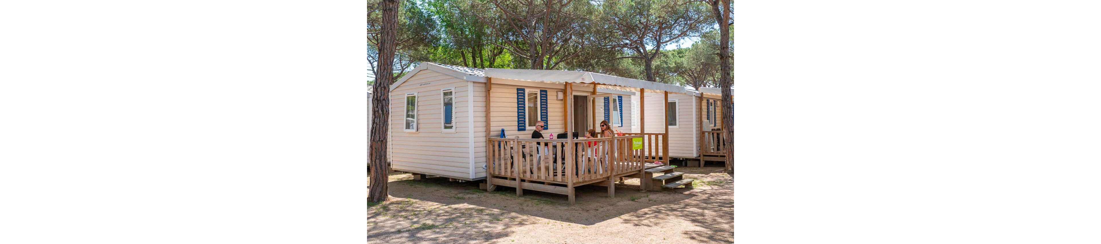 A family sits together on the wooden terrace of a light-colored mobile home. The cabin is set among tall pine trees in a sunny outdoor campground. Adults and children gather around a table, enjoying leisure time in the shade of the porch roof. The scene suggests a relaxed holiday atmosphere in a natural setting.