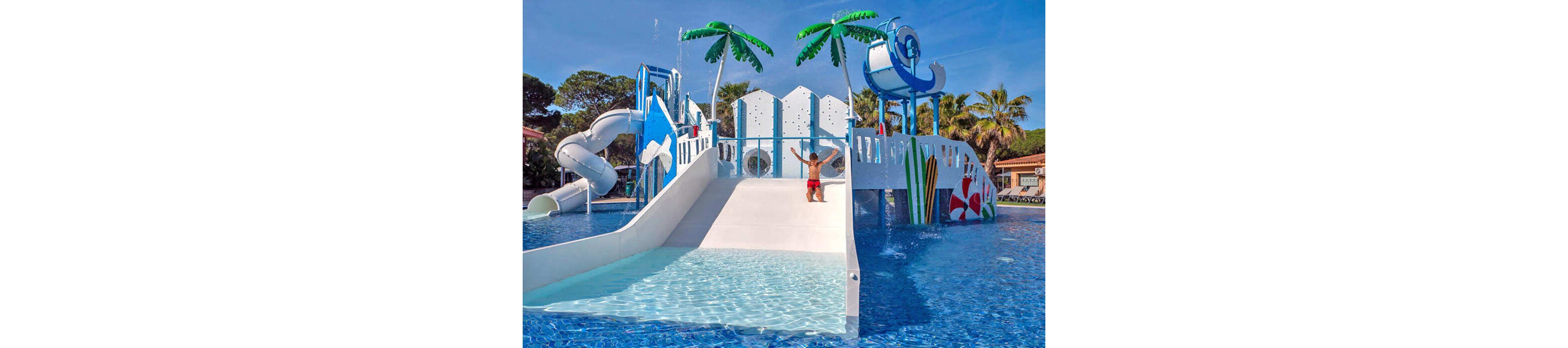 A young child slides down a wide white slide in a vibrant outdoor water park. The structure features multiple slides, climbing areas, and playful decorations in blue, white, and red. Palm tree sculptures and splashing water jets add to the tropical, fun atmosphere. Clear blue pool water surrounds the play area under a sunny sky.