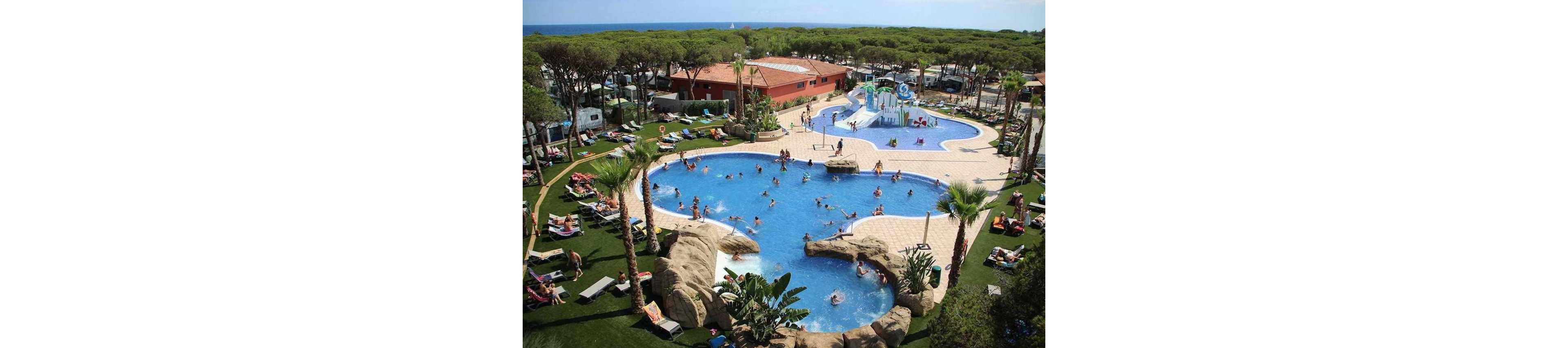 An outdoor resort features multiple interconnected swimming pools filled with people enjoying the water. Families and children play and swim while others relax on sun loungers around the poolside. The area is landscaped with rocks, palm trees, and green lawns, creating a tropical atmosphere. In the background, a water play area and slides are visible, with a forest of tall trees and the sea beyond.