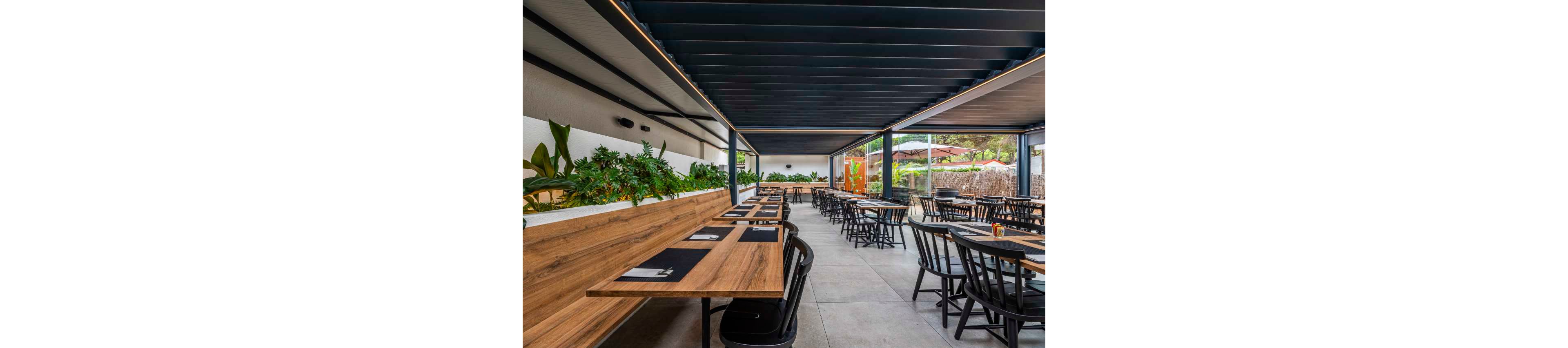 A contemporary open-air restaurant features long wooden tables and black chairs arranged in neat rows. The space is covered by a dark slatted roof with large open sides that reveal greenery and an outdoor view. Built-in planters with lush plants run along one side, adding a natural accent to the clean, minimalist design. The setting appears calm and ready for guests, with placemats and cutlery already laid out.