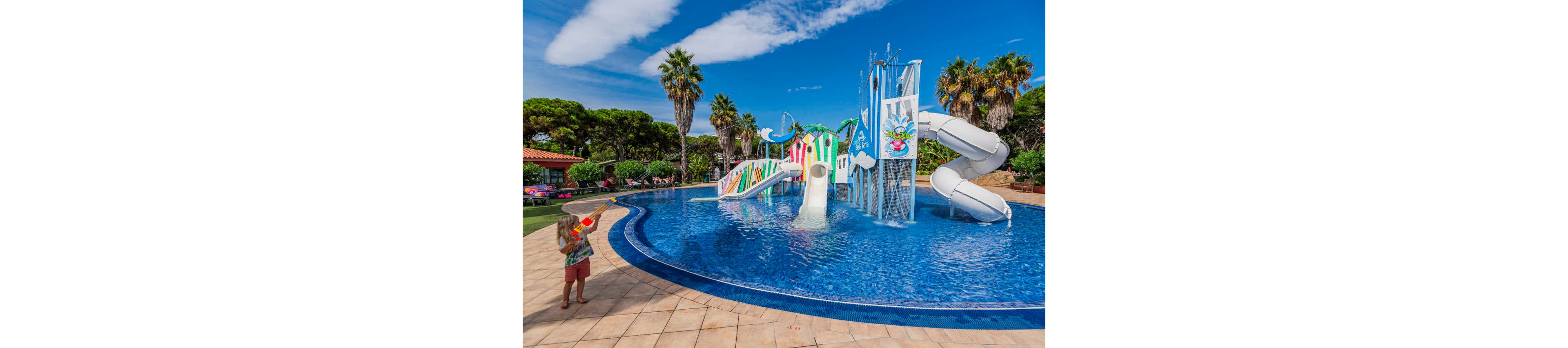 A young child stands at the edge of a large outdoor pool holding a water gun. In the background, a colorful water playground with slides and climbing structures rises from the center of the pool. Tall palm trees and lush greenery surround the area under a bright blue sky with scattered clouds. The scene conveys a fun, family-friendly vacation atmosphere at a sunny resort.
