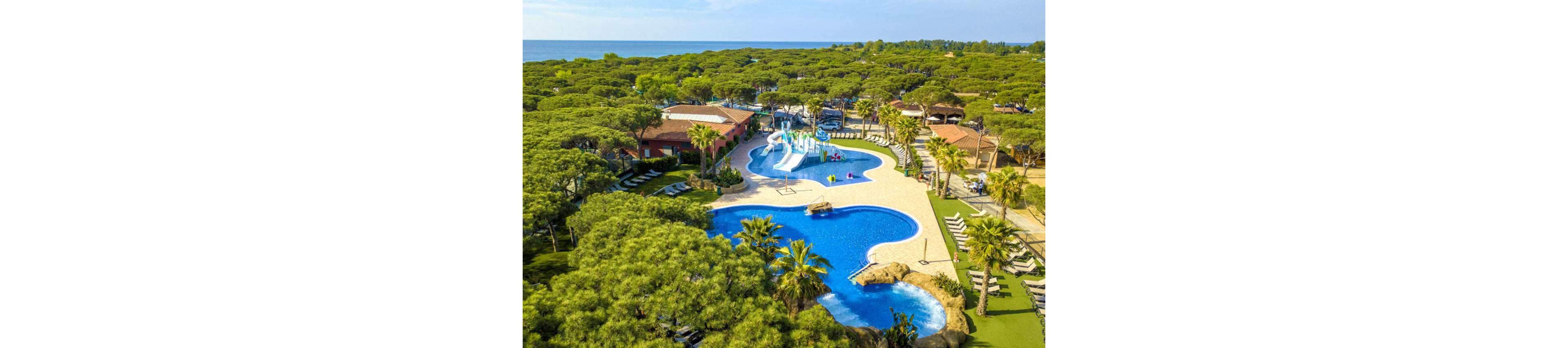 A large resort complex features multiple curving blue swimming pools surrounded by lush green trees and palm trees. In the center, a colorful water play area with slides and fountains is set up for children. Sun loungers and shaded seating areas line the poolside terraces, creating a relaxing vacation atmosphere. Beyond the resort, dense forest stretches toward the coastline and the distant sea.