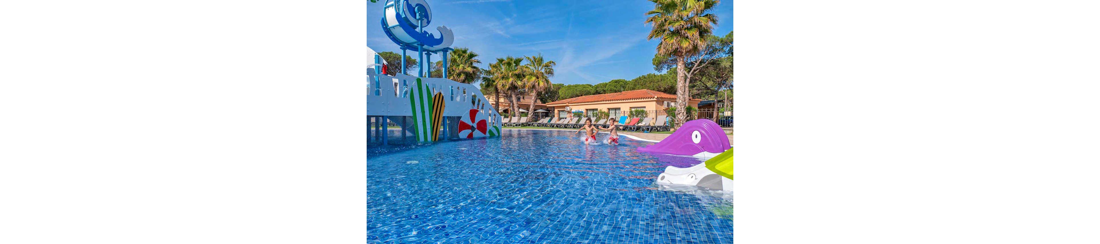 Two children play together in a shallow outdoor swimming pool at a sunny resort. The pool features a white play structure shaped like a ship with surfboard and candy decorations. Colorful animal slides and water toys are visible in the foreground, while palm trees and low buildings line the background. The clear blue sky and sparkling water create a bright, cheerful vacation atmosphere.