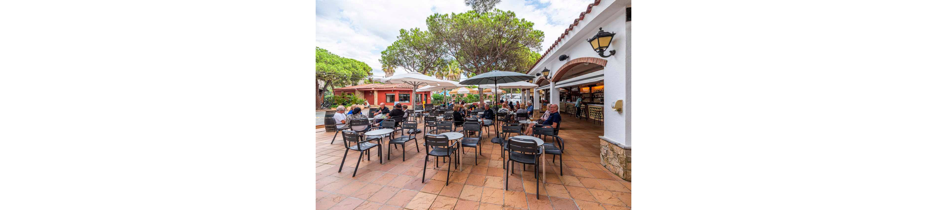 An outdoor café terrace is filled with people sitting at tables under large umbrellas. The scene is set beside a white stucco building with arched openings and warm wall lanterns. Guests appear relaxed, enjoying food and drinks in a casual social atmosphere. Trees and additional buildings frame the open tiled patio, suggesting a resort or holiday setting.