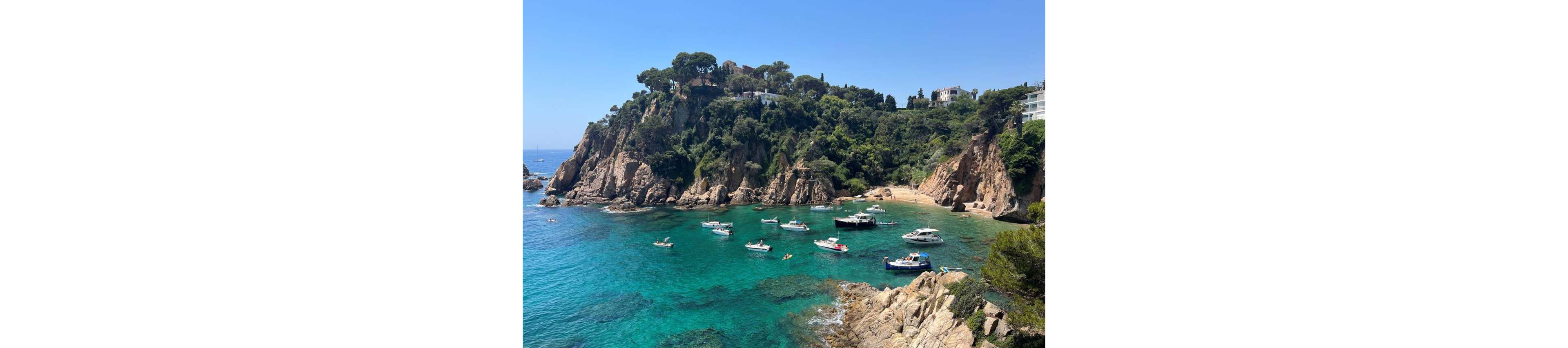 Small boats float on clear turquoise water in a sheltered rocky cove. A lush, tree-covered cliff rises above the bay, with a few white houses perched along the hillside. People relax on the boats and swim near the shore under a bright blue sky. The scene evokes a peaceful summer day on a picturesque Mediterranean coastline.