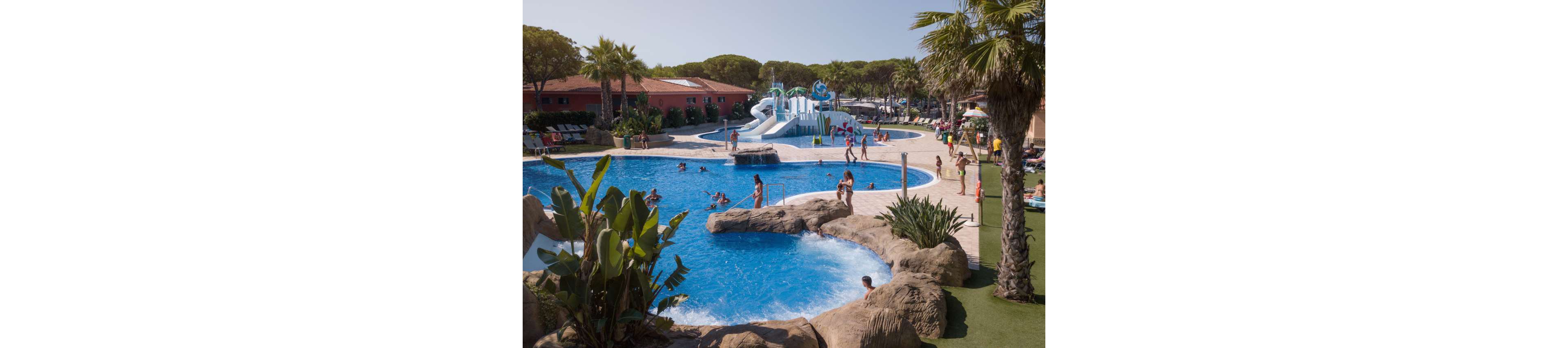 An outdoor resort swimming pool is filled with families and children playing in the water. A large white and blue water slide structure rises in the background, with several people climbing the stairs and sliding down. Palm trees, tropical plants, and artificial rock formations surround the pool, creating a vacation atmosphere. Sun loungers and umbrellas line the far side, suggesting a relaxed holiday setting under clear skies.