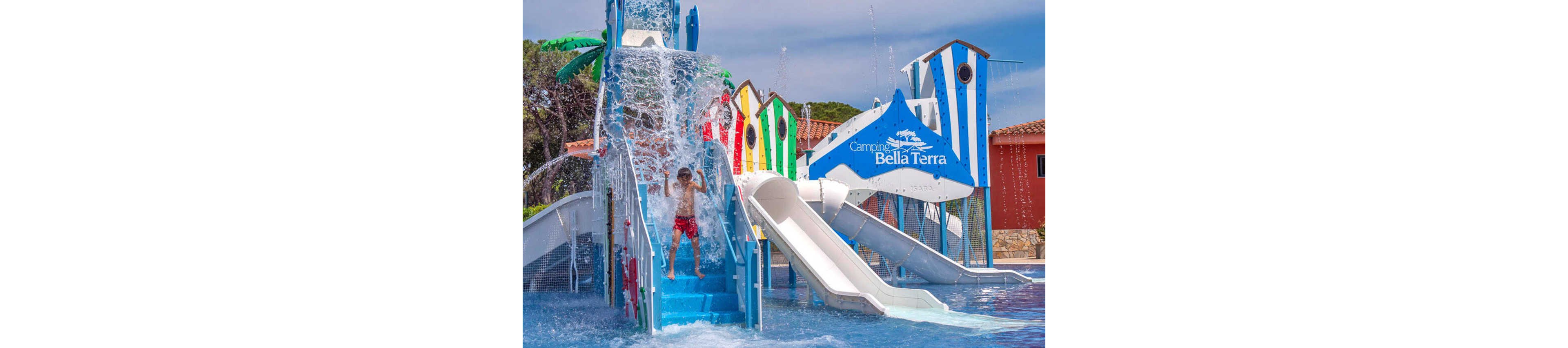 A young boy stands under a splash of water on a bright blue staircase at an outdoor water park. Colorful striped structures and multiple slides surround the shallow pool area. The sign "Camping Bella Terra" is clearly visible on a large decorative element above the slides. The scene suggests a fun summer day at a family-friendly campsite or resort.
