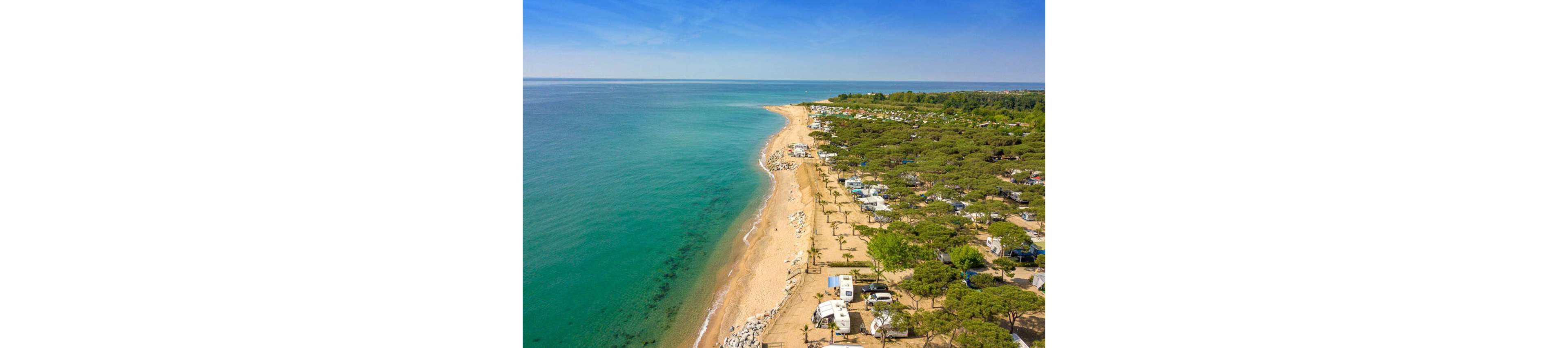 An aerial view shows a long sandy beach bordering a calm turquoise sea. A large campground with caravans, RVs, and tents stretches along the shoreline beneath dense green trees. The coastline curves gently into the distance under a clear blue sky. The scene suggests a peaceful seaside holiday destination with direct beach access.