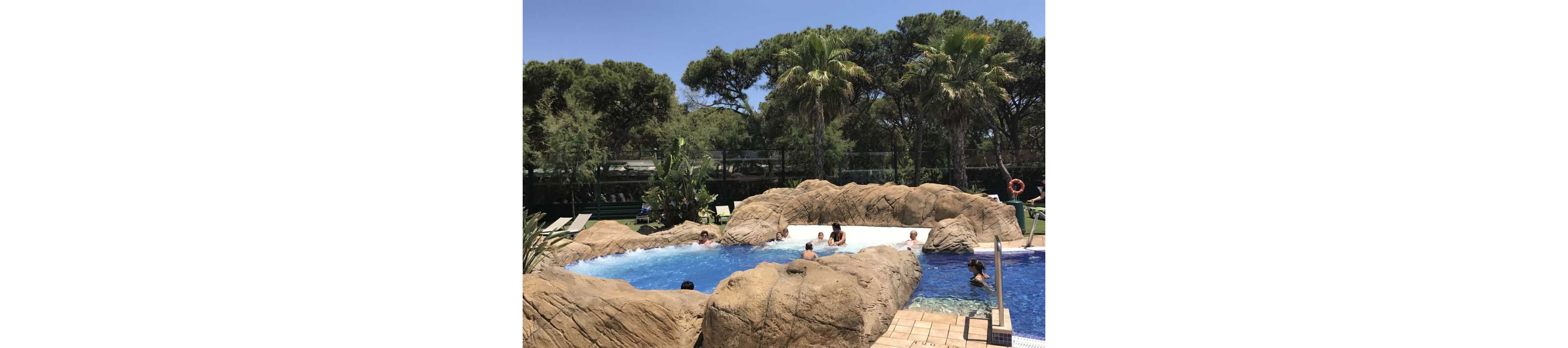 A group of people relax and play in a resort-style swimming pool surrounded by artificial rock formations. The pool has different water levels, with adults and children enjoying the shallow and deeper sections. Palm trees and dense green foliage create a tropical atmosphere under a clear blue sky. Sun loungers and poolside tiles frame the scene, suggesting a warm vacation day outdoors.