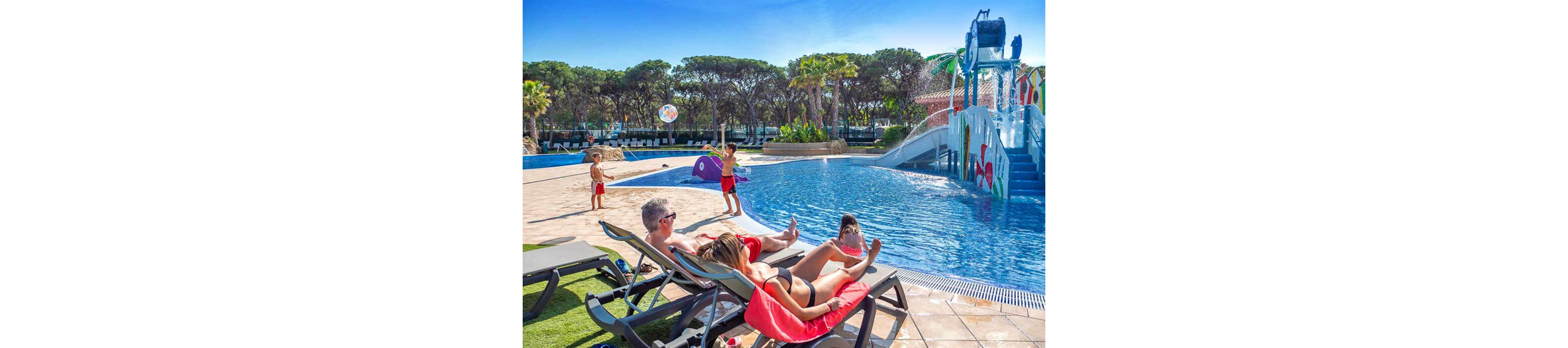 Adults lounge on sunbeds beside a bright blue resort swimming pool while children play nearby. A colorful water play structure with slides and fountains rises from the pool in the background. Tall trees and palm trees surround the area under a clear blue sky, creating a holiday atmosphere. The scene suggests a warm, sunny day at a family-friendly vacation destination.